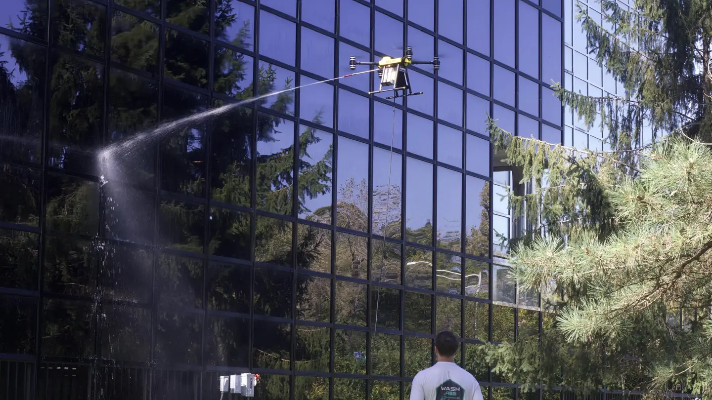 A drone sprays water on the glass wall of an office building while a pilot watches from the ground