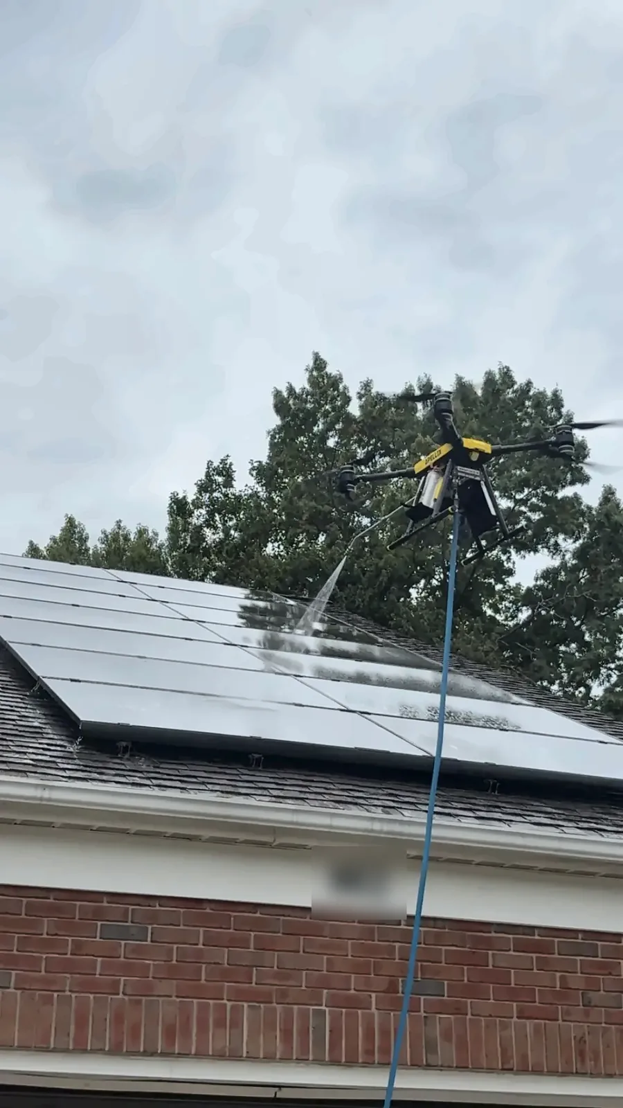 A tethered drone rinses the lower row of a solar array while its water hose runs down to the ground