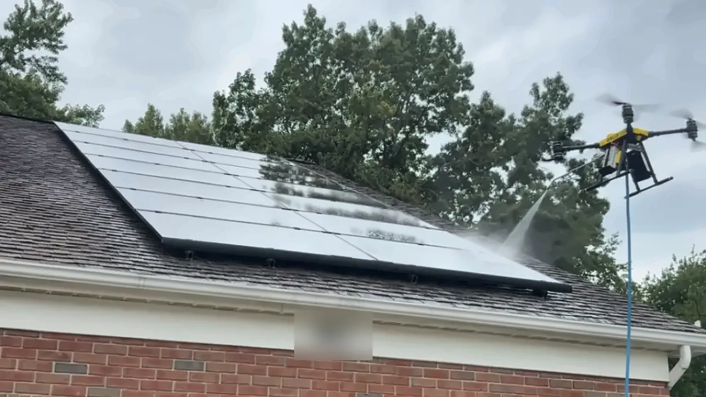 A cleaning drone sprays a rooftop solar array on a garage, with tall trees behind the roof